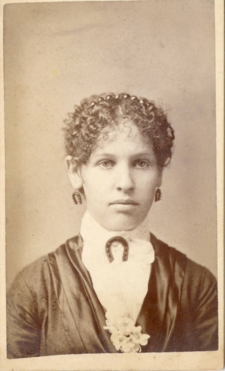 Black and white carte de visite photograph of a woman with short, dark hair who wears a horseshoe broach and matching earrings.